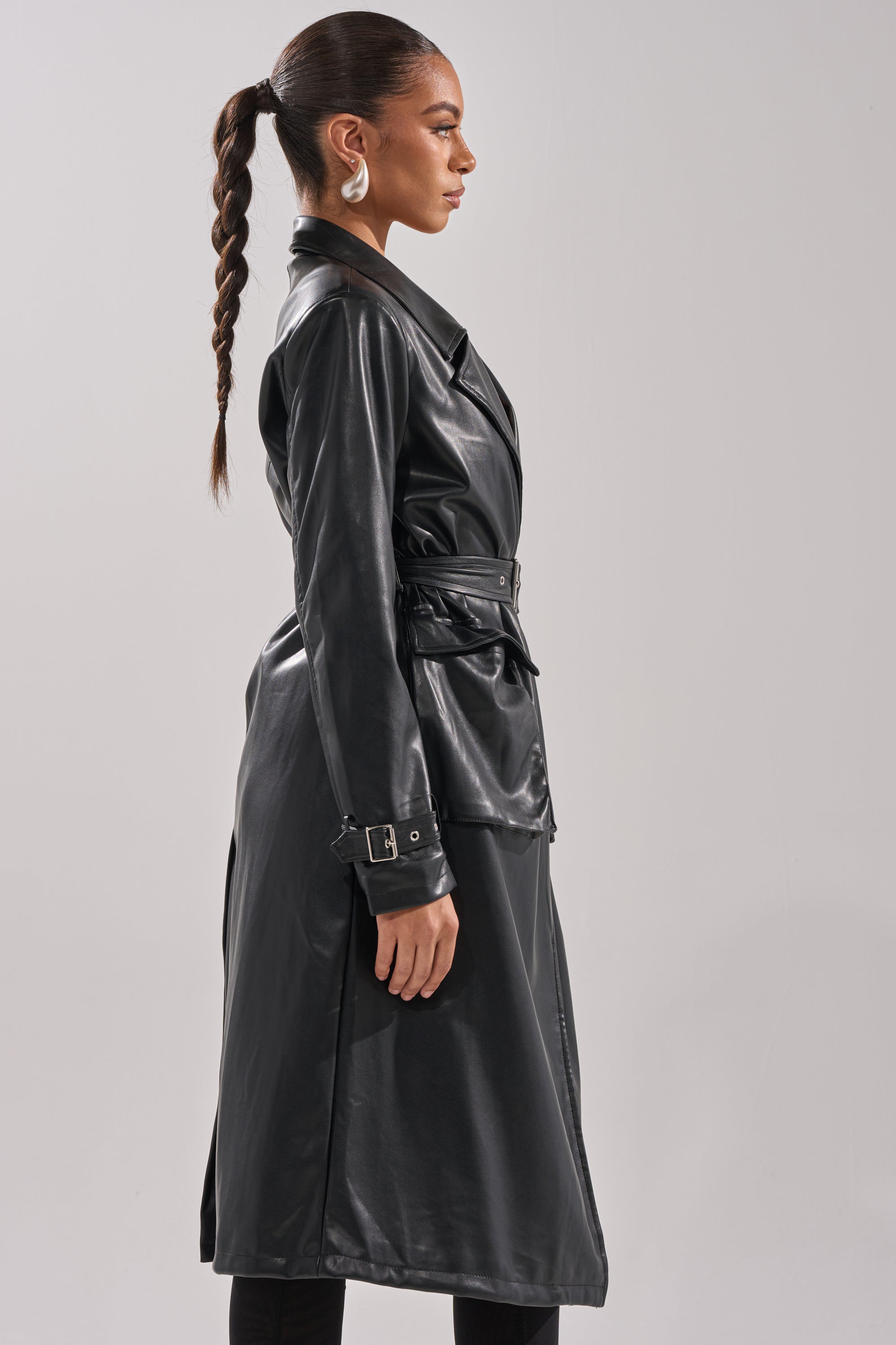 A woman with a long braided ponytail wears the MONTANNA BUTTER LUXE EVERYDAY TRENCH and large white earrings, standing in profile against a plain light gray background. 3