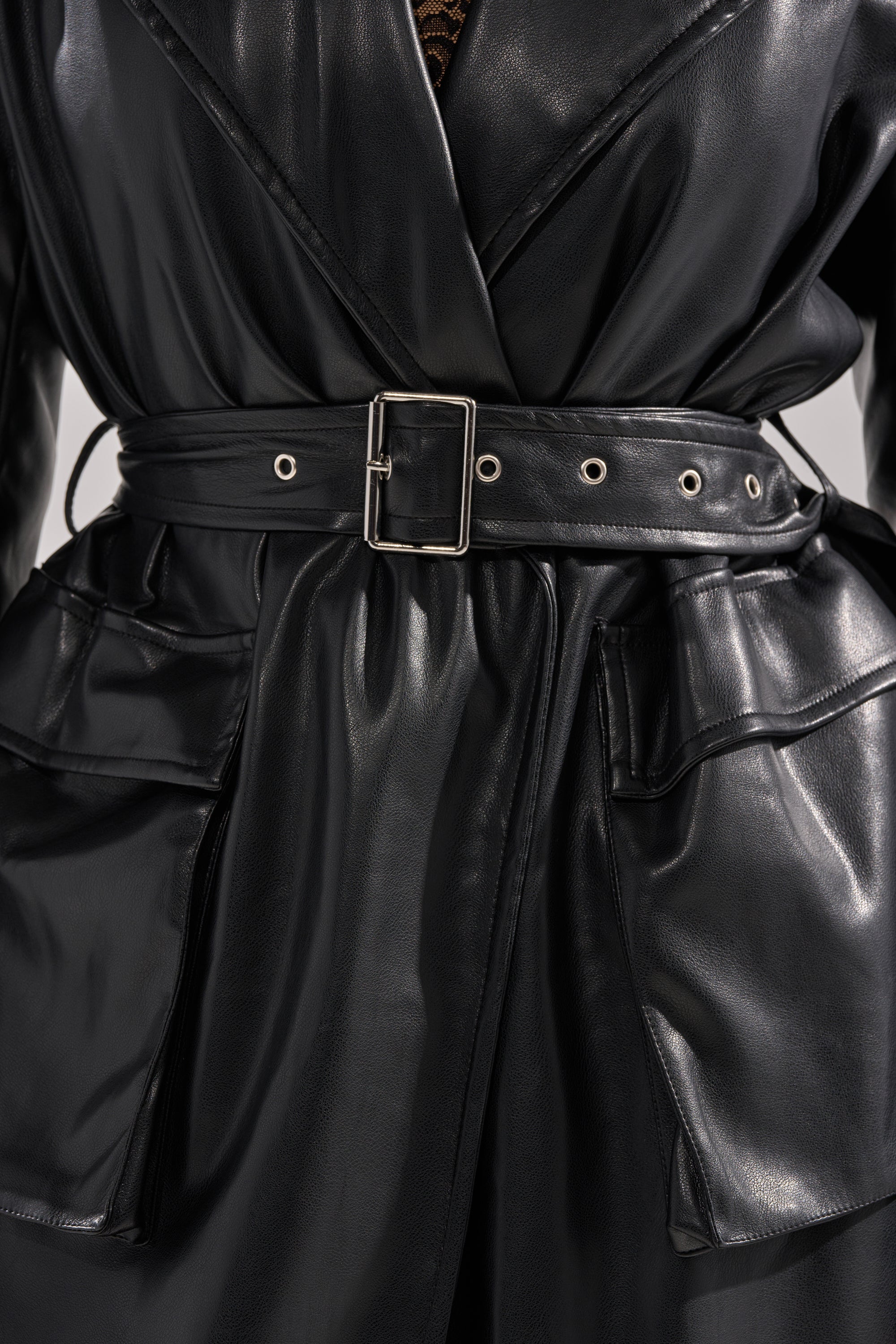 Close-up of a person wearing the MONTANNA BUTTER LUXE EVERYDAY TRENCH in shiny black vegan leather, featuring a wide belt with a silver rectangular buckle and large pockets. 6