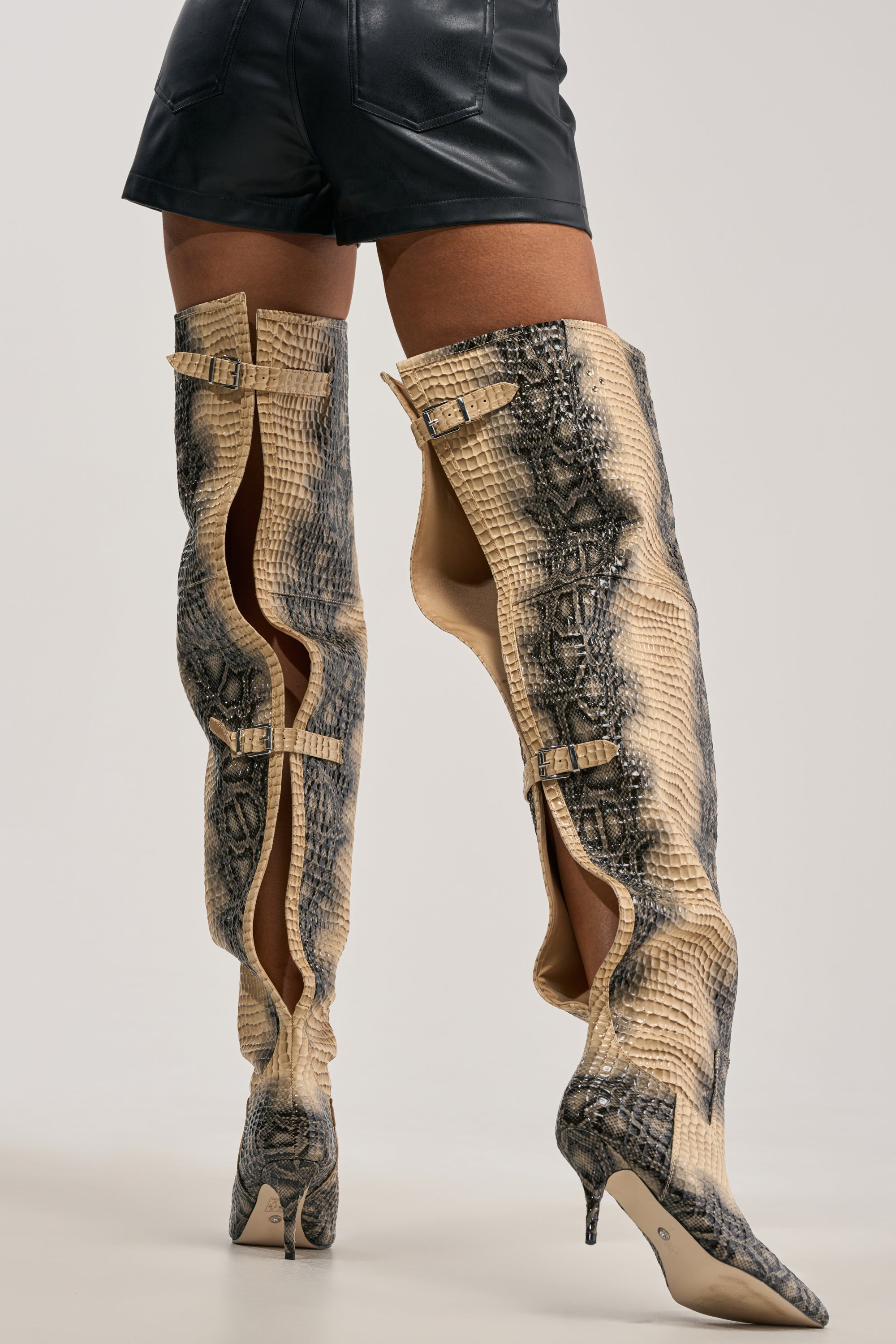 Wearing black leather shorts and the AZALEA WANG MOONREEL SNAKE THIGH HIGH BOOT—tall, snake print stiletto boots with buckles—a person stands against a plain background, exuding bold streetwear style.
