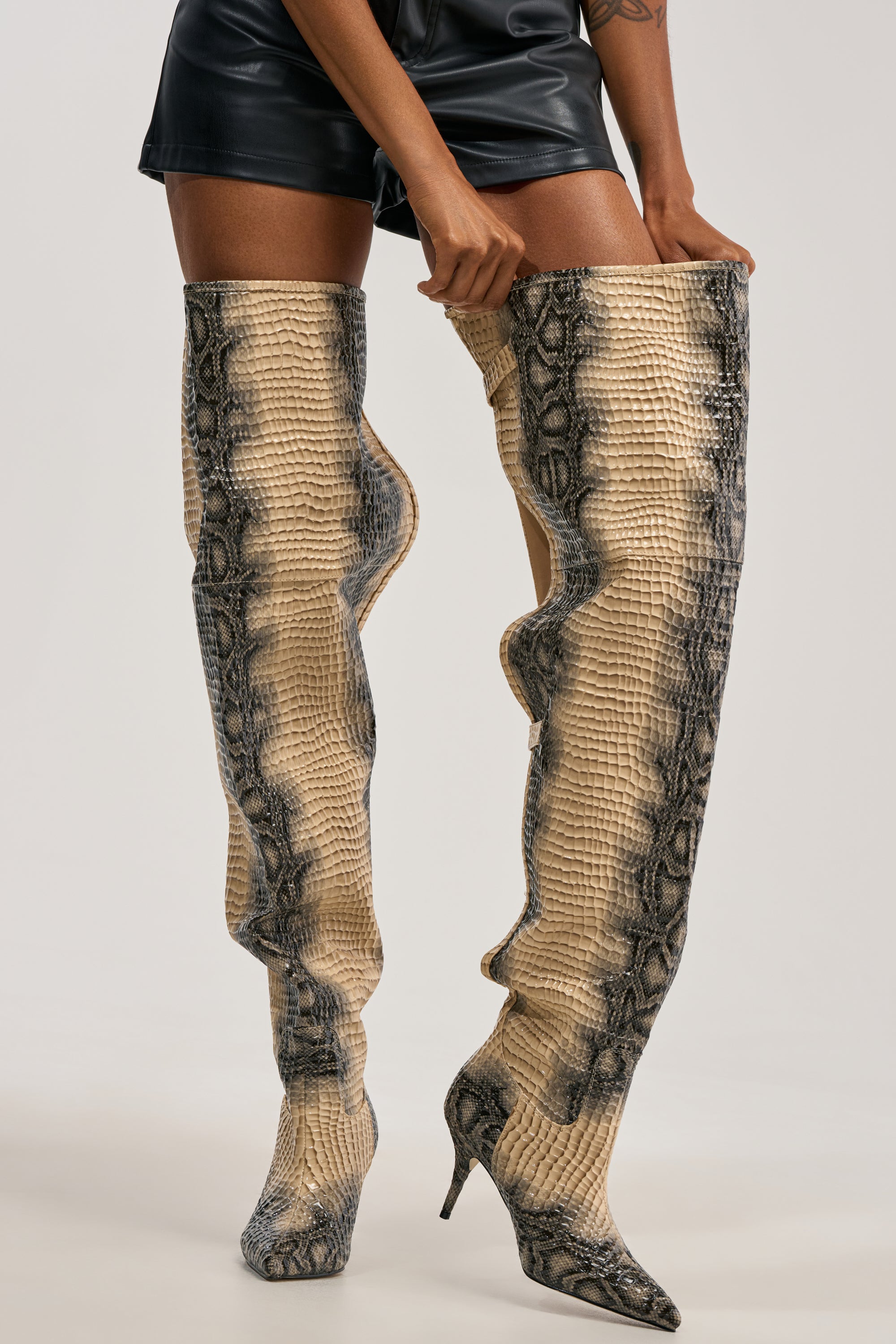 Wearing black leather shorts and the AZALEA WANG MOONREEL SNAKE THIGH HIGH BOOT—tall, slouchy stiletto boots with a striking snakeskin pattern—a person holds the top edges of the boots for an edgy streetwear vibe.