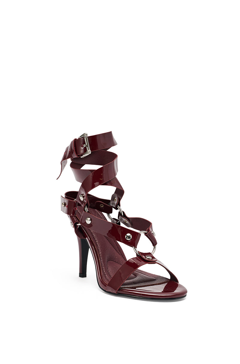 The AZALEA WANG MORGANNA Burgundy Patent Sexy Stiletto Sandal features wide crisscross patent leather straps, metal studs, and a silver-buckled ankle strap, shown against a white background.