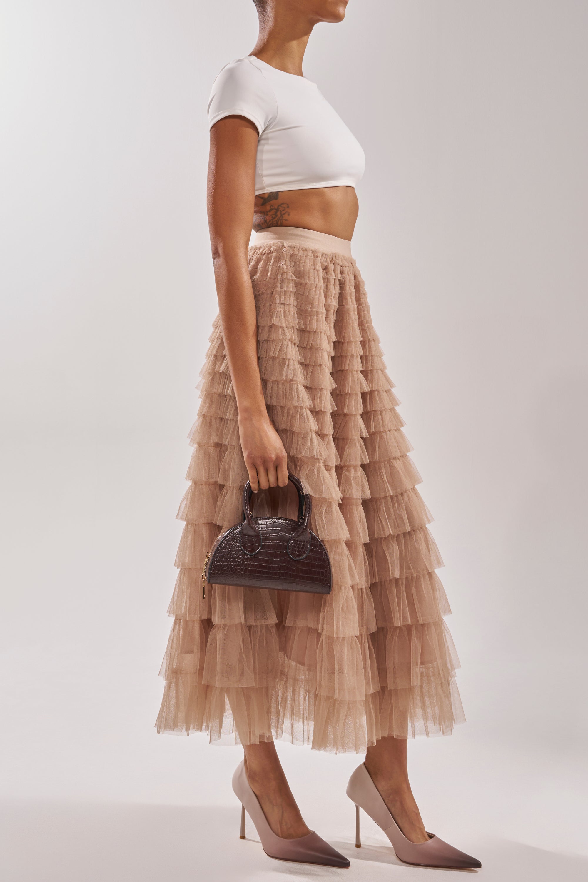 A woman wears a white crop top, a layered beige tulle midi skirt, the AZALEA WANG BROWN OMBRE EFFECT STILETTO PUMP, and holds a small dark brown handbag while standing against a plain light background. 1