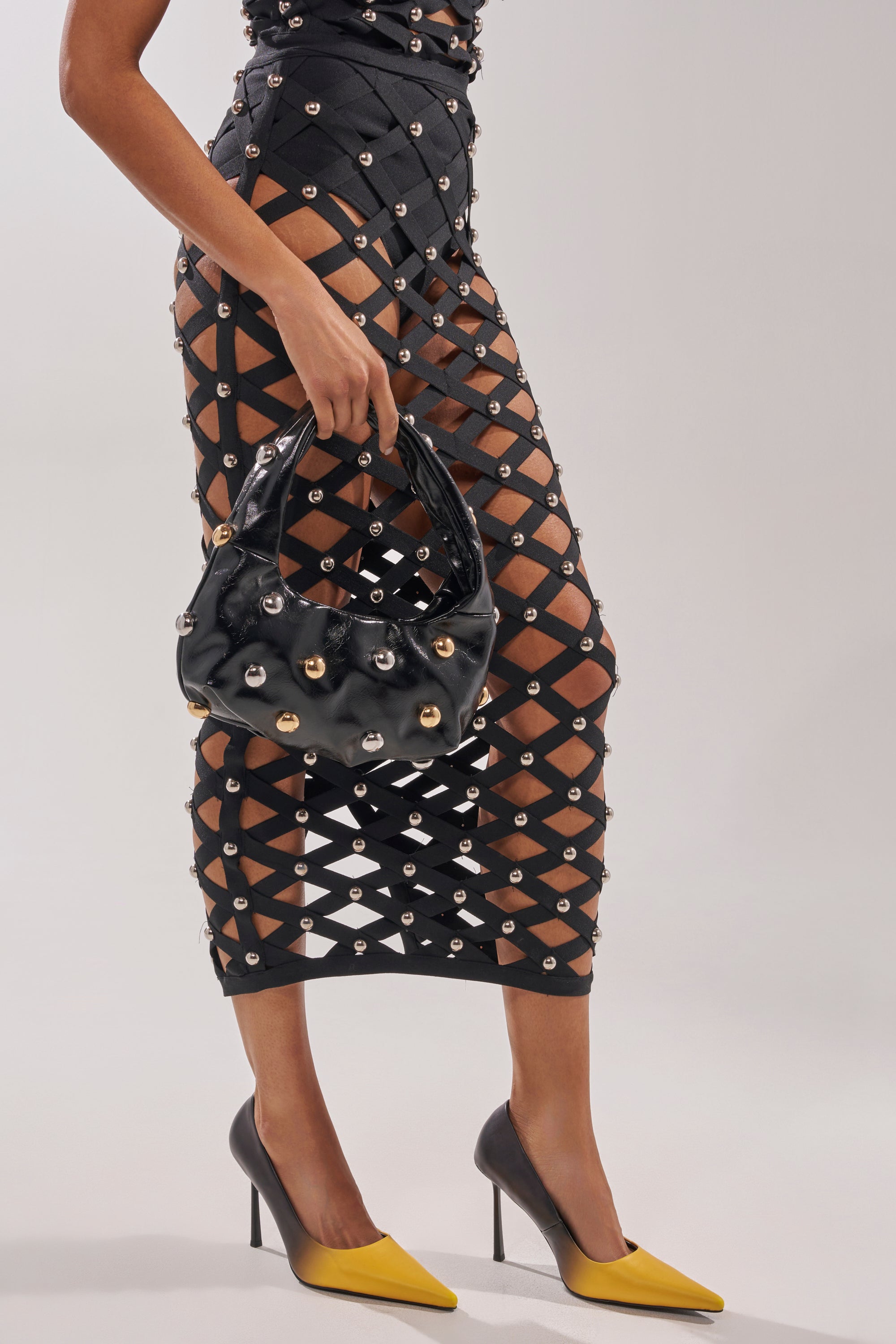 A person wears a black lattice skirt with metallic grommets, carries a matching faux leather handbag, and completes the look with AZALEA WANG YELLOW OMBRE EFFECT STILETTO PUMPS. The background is plain and light. 1