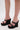 A person stands on a light surface wearing AZALEA WANG MORITZ BLACK SPIKEY WEDGE SANDALS. The ankle straps, platform sole, and small spikes create a bold punk-glam style.
