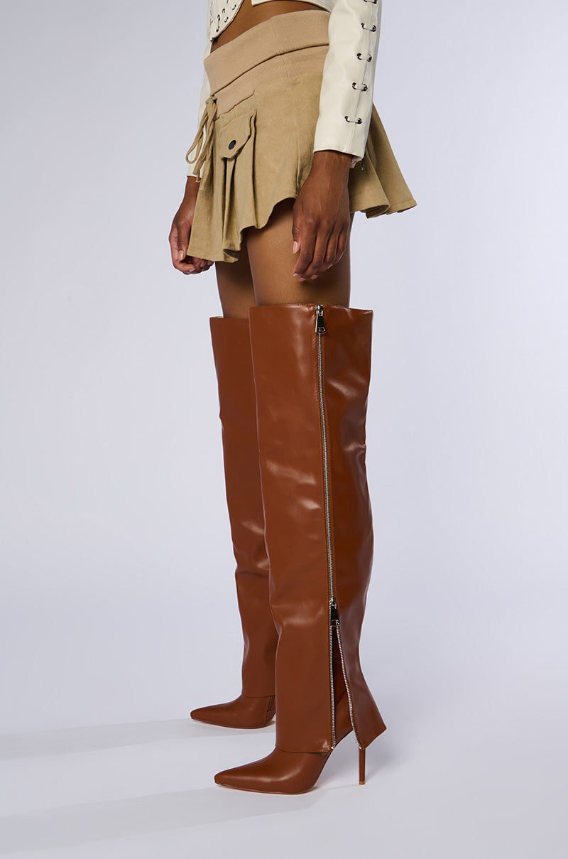 A person wearing a beige mini skirt, beige jacket, and the stylish AZALEA WANG MOSSY TAN BOOTS with side zippers stands against a plain background. Their hands rest casually by their sides, and their outfit exudes contemporary chic. 1