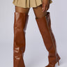 A person wearing a beige mini skirt and the AZALEA WANG MOSSY TAN BOOT, a pair of high-heeled, thigh-high brown leather boots with a side zipper. The background is plain and light-colored. The outfit emphasizes the boots and skirt, showcasing a stylish and bold fashion choice.