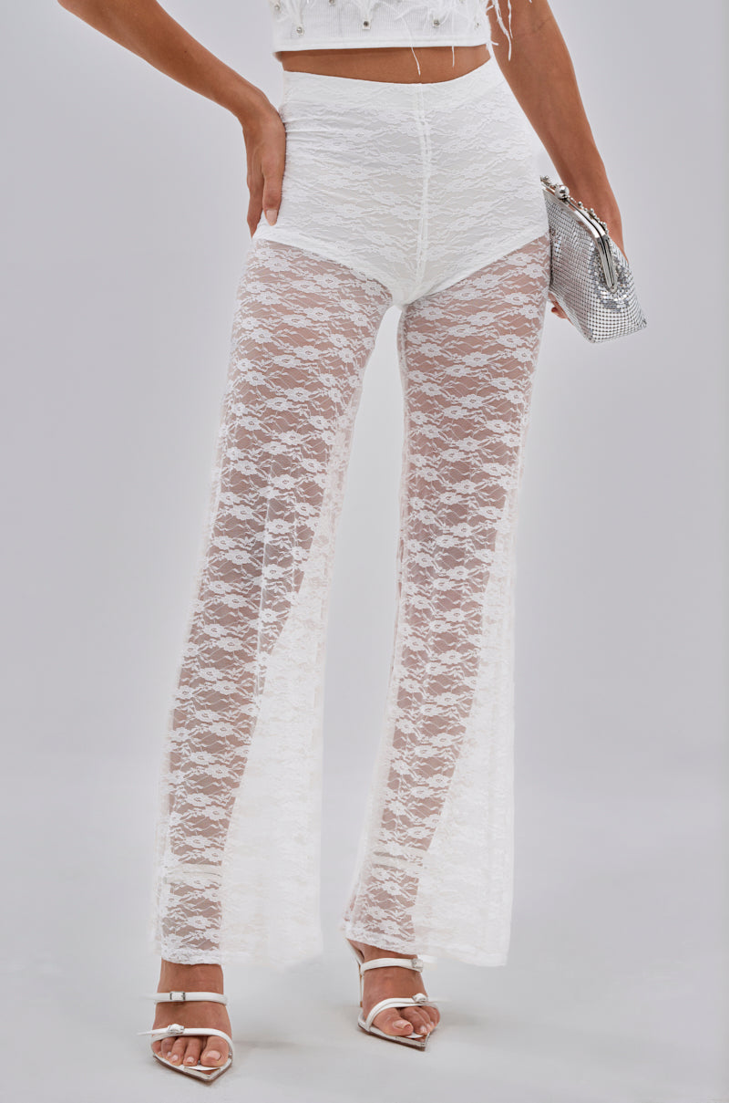 A person wears the HOT STUFF LACE FLARE LEGGING, a sheer floral lace, high-waisted festival bottom, paired with a matching white lace crop top, white high-heeled sandals, and a silver clutch purse. 0