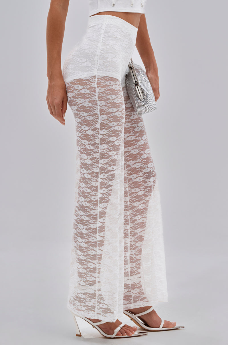 Wearing the HOT STUFF LACE FLARE LEGGING in sheer floral white, paired with a white crop top, white heels, and holding a silver textured clutch bag, a person stands against a plain light gray background. 2