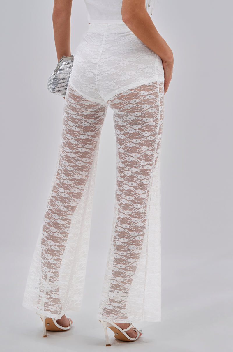 Wearing HOT STUFF LACE FLARE LEGGING over white underwear and high-heeled sandals, holding a small silver clutch, this back view highlights the elegant, sheer floral festival style. 3
