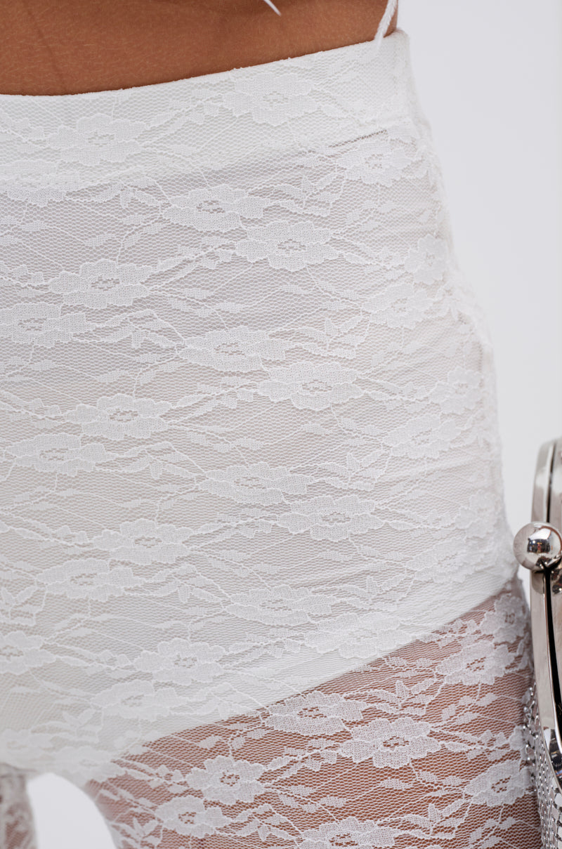 Close-up of a person wearing the HOT STUFF LACE FLARE LEGGING—high-waisted, white, sheer floral lace bottoms. A silver purse is partially visible to the right, highlighting the elegant and delicate lace pattern. 7