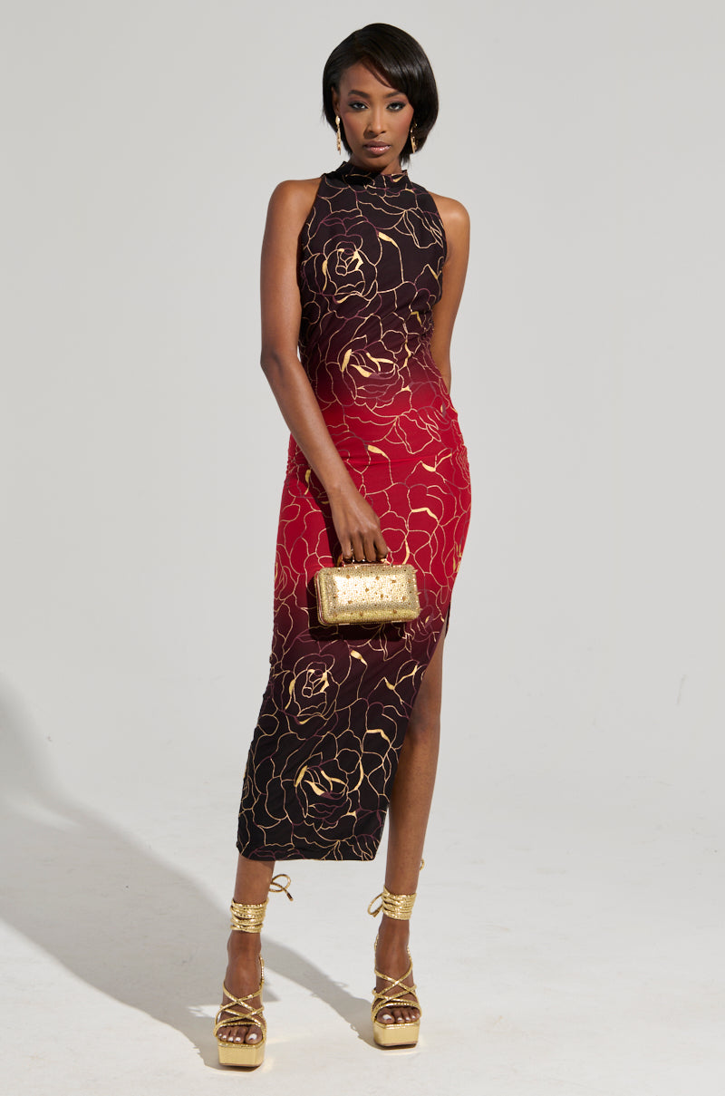 A person poses confidently in a sleeveless, red and black IRIS METALLIC OMBRE PRINTED MIDI DRESS with a high neckline and side slit. They hold a gold clutch and wear gold platform heels, standing against a plain gray background.