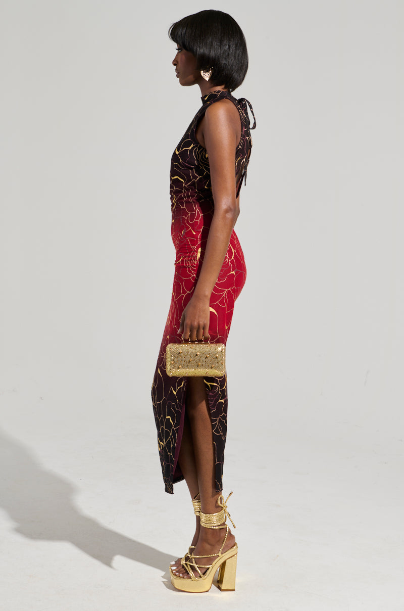 A woman in the IRIS METALLIC OMBRE PRINTED MIDI DRESS, characterized by its body-hugging, sleeveless design with a floral outline pattern in shades of black and red. She is accessorizing with a gold clutch, complemented by gold high-heeled sandals and large earrings, as she poses against a plain background.