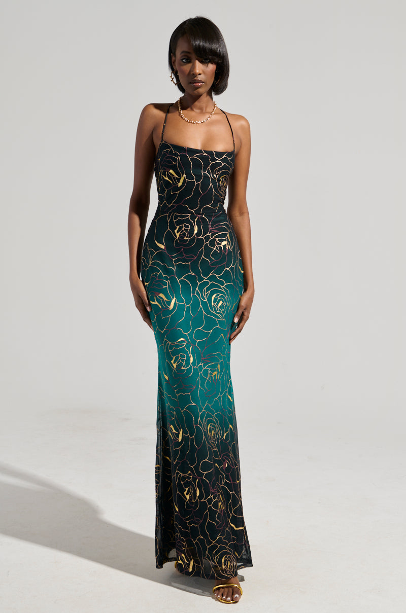 Against a plain gray background, a person confidently stands in the LUNA METALLIC OMBRE PRINTED MIDI DRESS, which showcases a stunning mermaid silhouette with a gradient from black to teal and is adorned with an exquisite gold floral pattern. The dress has thin straps and is perfectly accessorized with a necklace and sandals.