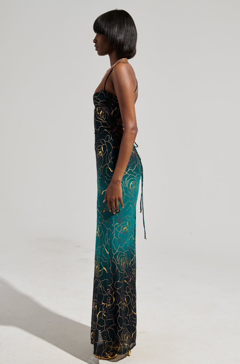 A person with shoulder-length dark hair stands in profile, wearing the fitted LUNA METALLIC OMBRE PRINTED MIDI DRESS featuring thin straps and a shimmering gold design. This dress elegantly highlights their form against a neutral gray background.