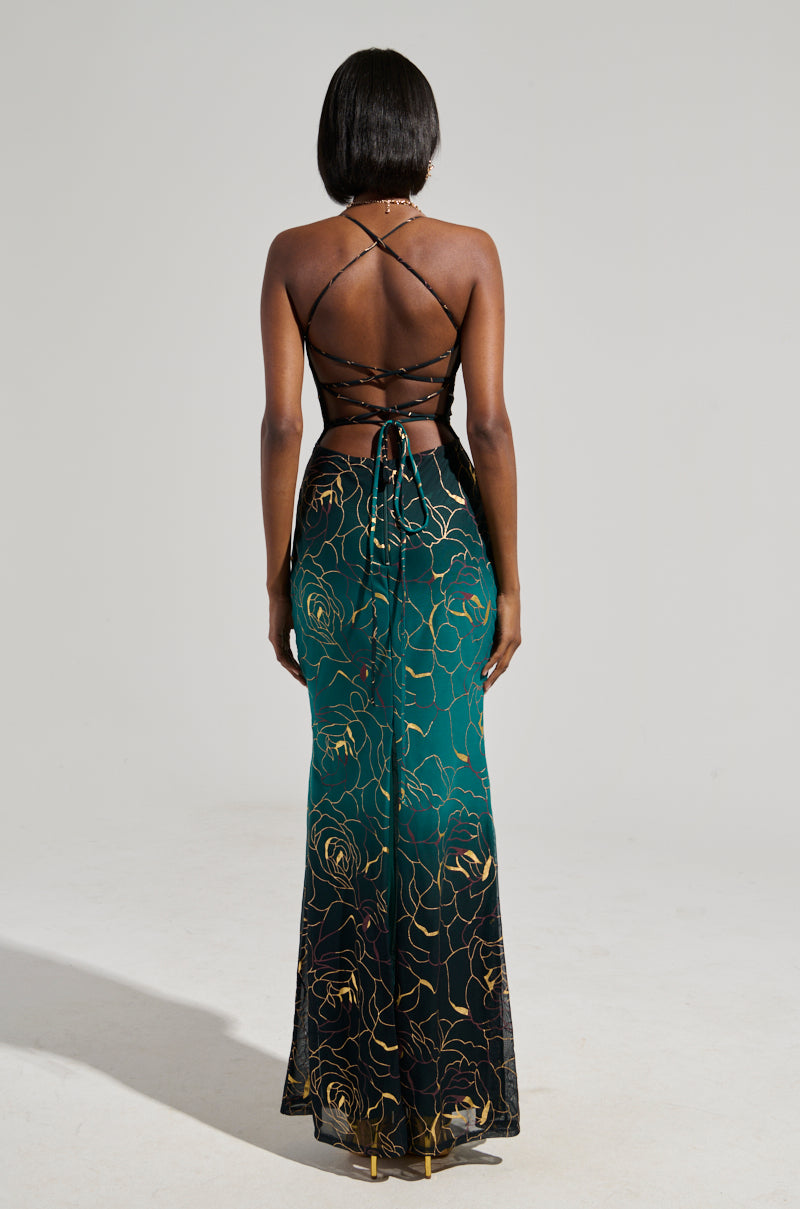 A woman with shoulder-length hair is wearing a stunning Luna Metallic Ombre Printed Midi Dress, which boasts an intricate strap design on the back. The dress features a gradient from teal to black with a captivating floral pattern. She stands facing away from the camera, exuding elegance and grace.