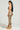 A woman with long dark hair stands in profile, wearing the YOUR NEW OBSESSION ABSTRACT MIDI DRESS. She complements her outfit with gold high-heeled platform sandals and large hoop earrings as she faces left against a plain neutral background.