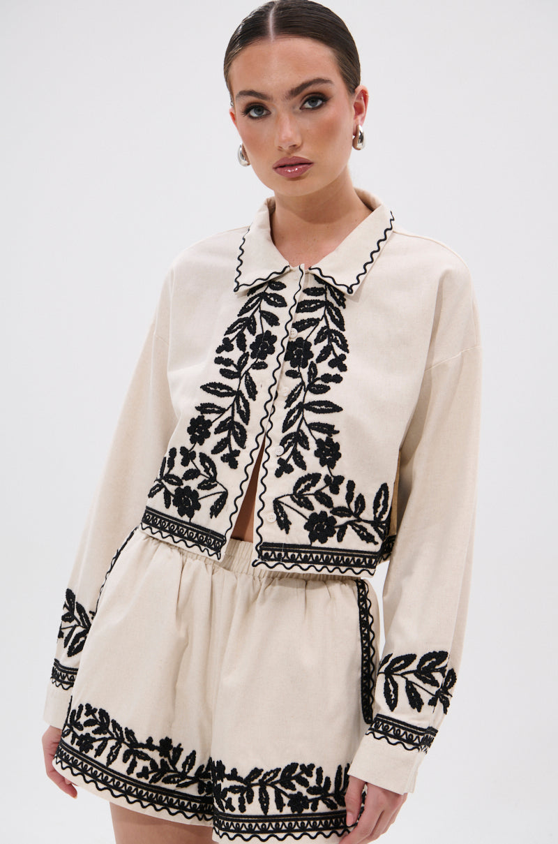 A woman poses in the FOREVER AND EVER FLORAL BUTTON DOWN—a cream lightweight linen top with black floral embroidery on the collar, front, cuffs, and hem. She styles it with slicked-back hair, natural makeup, and silver hoop earrings.