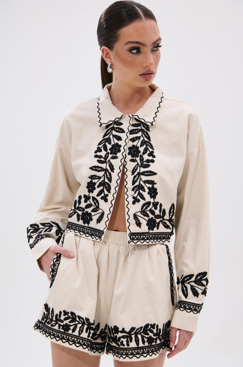 A woman wears the FOREVER AND EVER FLORAL BUTTON DOWN, a beige linen two-piece set with a cropped long-sleeve jacket and matching shorts, both detailed with black floral patterns and scalloped edges. She has sleek hair and looks to the side.