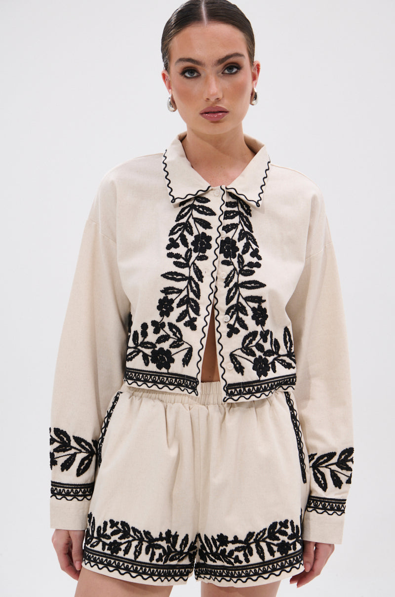 A woman wears the FOREVER AND EVER FLORAL BUTTON DOWN, a cream long-sleeve, collared bohemian top with black floral embroidery and wavy trim, paired with matching shorts. She looks ahead neutrally against a plain white background.