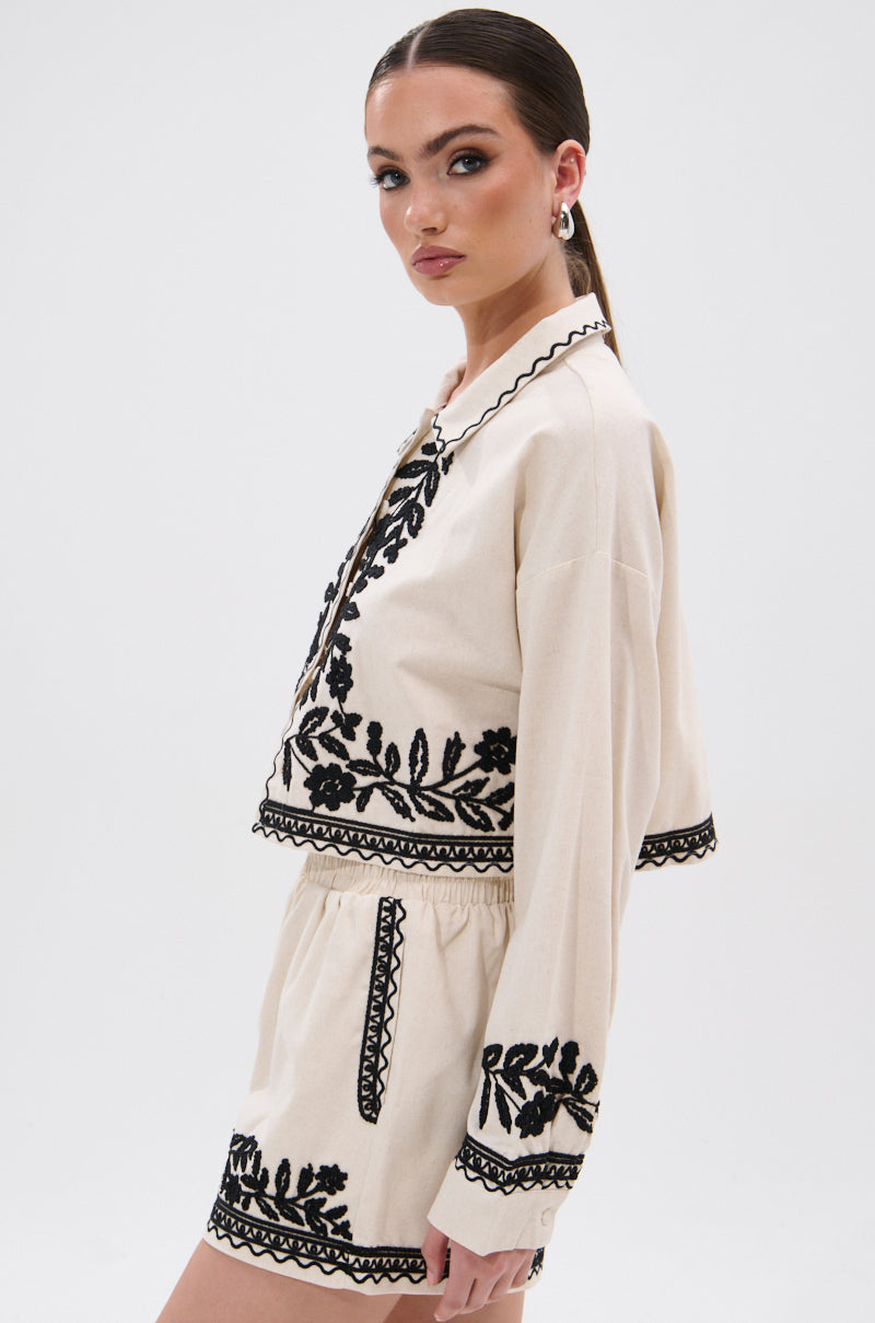 A woman stands against a plain white background, looking over her shoulder at the camera and wearing the FOREVER AND EVER FLORAL BUTTON DOWN, a cream-colored top with black floral embroidery in a bohemian style.