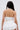 A person with long dark hair, seen from behind, wears the LUCY WHITE CORSET with lace-trimmed straps and a layered white skirt. A small tattoo appears on their left arm. The plain background is light-colored.