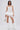 A woman stands confidently in the LUCY WHITE CORSET with lace-trimmed straps, styled with an asymmetrical ruffled mini skirt, white knee-high boots, layered necklaces, and a metallic purse against a plain white background.