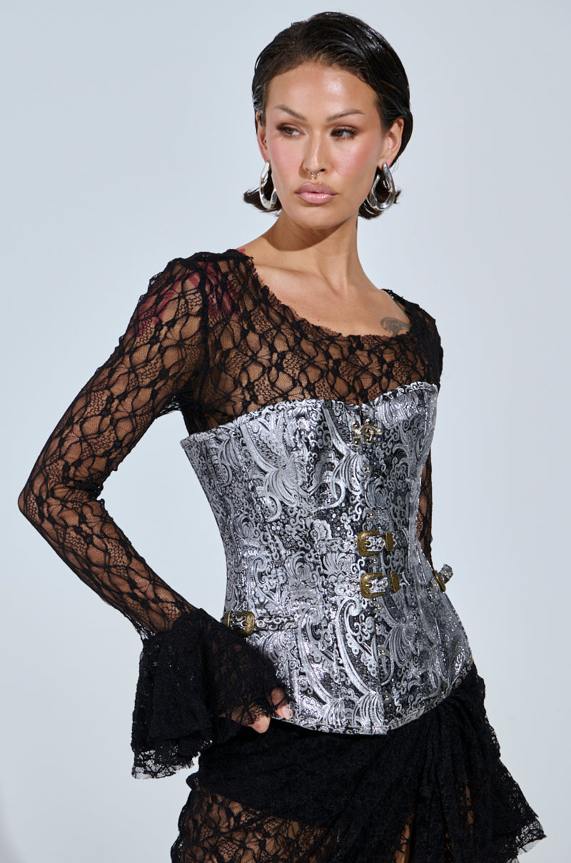 A woman with slicked-back hair and silver hoop earrings wears a sheer black lace top with flared sleeves, the SILVER PAISLEY CORSET TOP with metallic hook closures, and a black textured skirt, looking to the side.