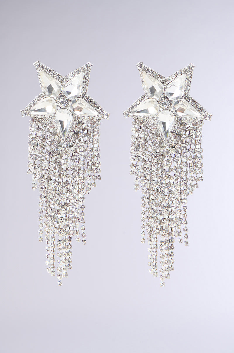 The SHOOTING STAR BLING EARRING showcases an ornate design with large, star-shaped features encrusted with clear rhinestone embellishments. Multiple strands dangle beneath the stars, adorned with smaller sparkling crystals that create a cascading and shimmering effect against a light gray background.
