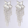 The SHOOTING STAR BLING EARRING showcases an ornate design with large, star-shaped features encrusted with clear rhinestone embellishments. Multiple strands dangle beneath the stars, adorned with smaller sparkling crystals that create a cascading and shimmering effect against a light gray background.