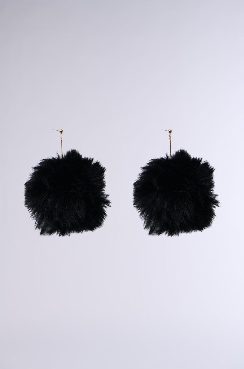 The TUNDRA FAUX FUR EARRINGS showcase large, plush black pom-poms on thin gold hooks, set against a gray background. These fluffy faux fur earrings are the perfect statement makers for any accessory collection.