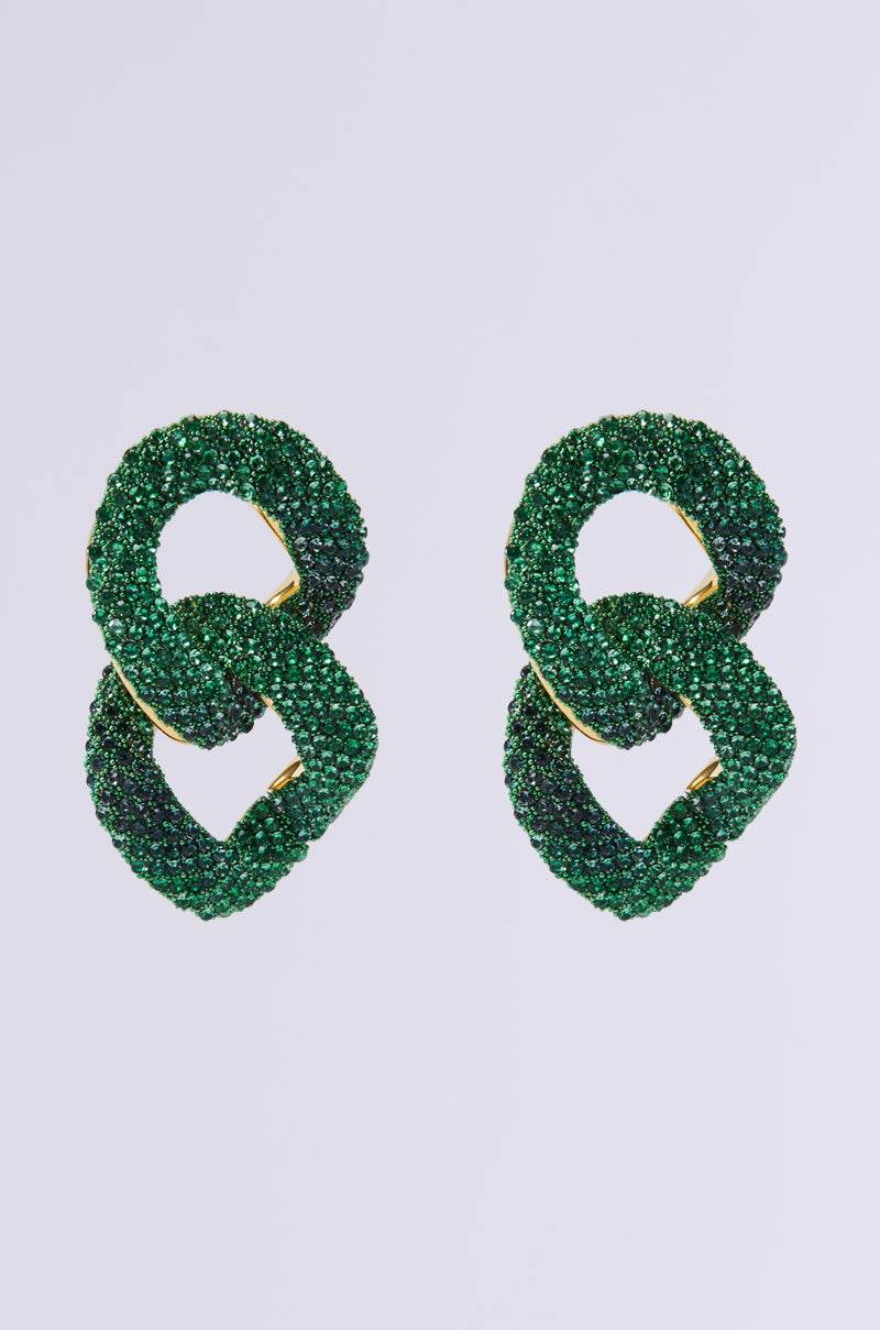 A pair of THE VISION RHINESTONE EARRINGS adorned with vibrant green crystals. These earrings are crafted on a metallic base and designed to interlock in an infinity shape, standing out against the simple light gray background.