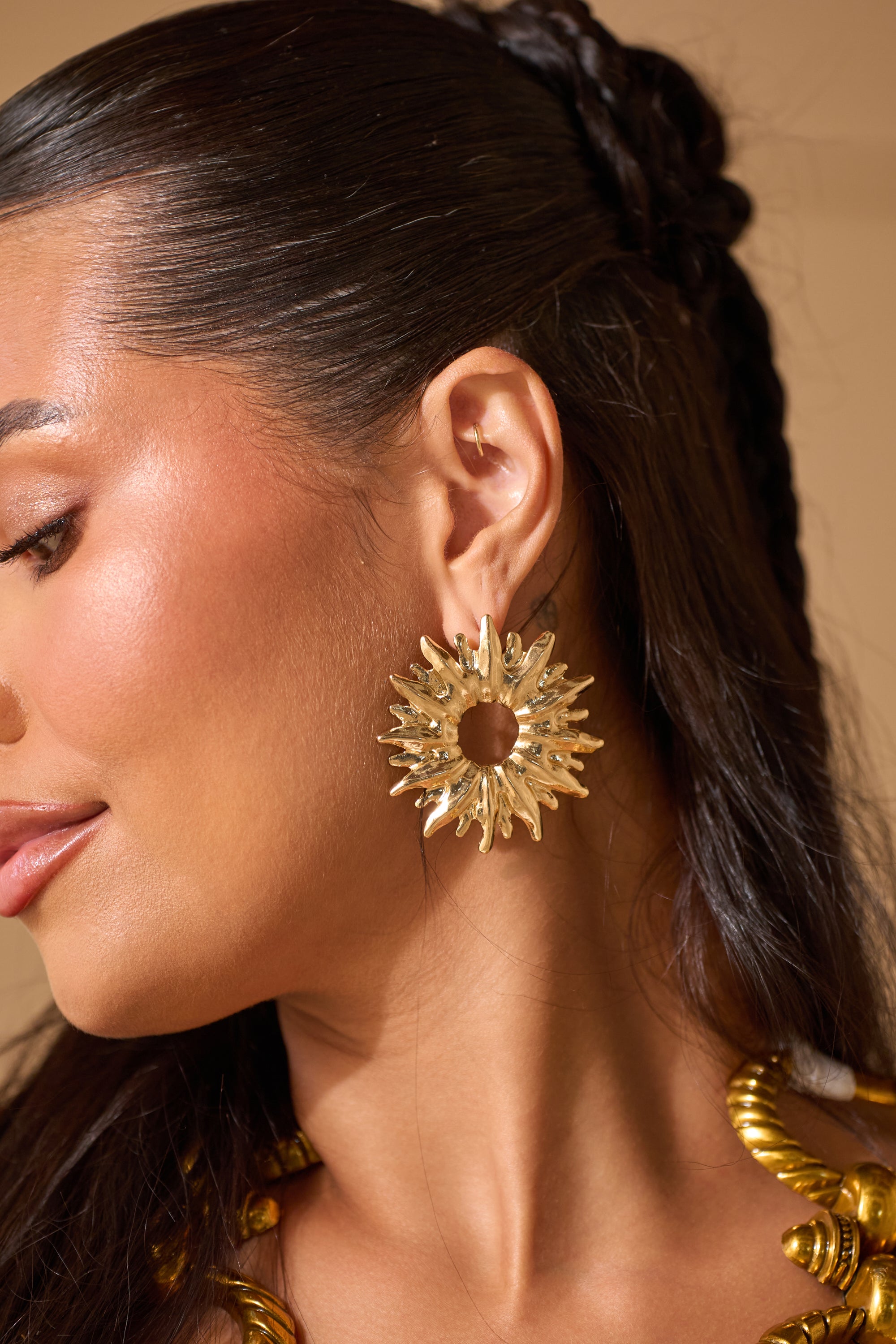 A woman with long, dark braids wears bold SUNBURST EARRING and a chunky gold necklace. The photo highlights her profile and jewelry, making the SUNBURST EARRING a stunning wedding guest accessory that complements glowing skin. 0