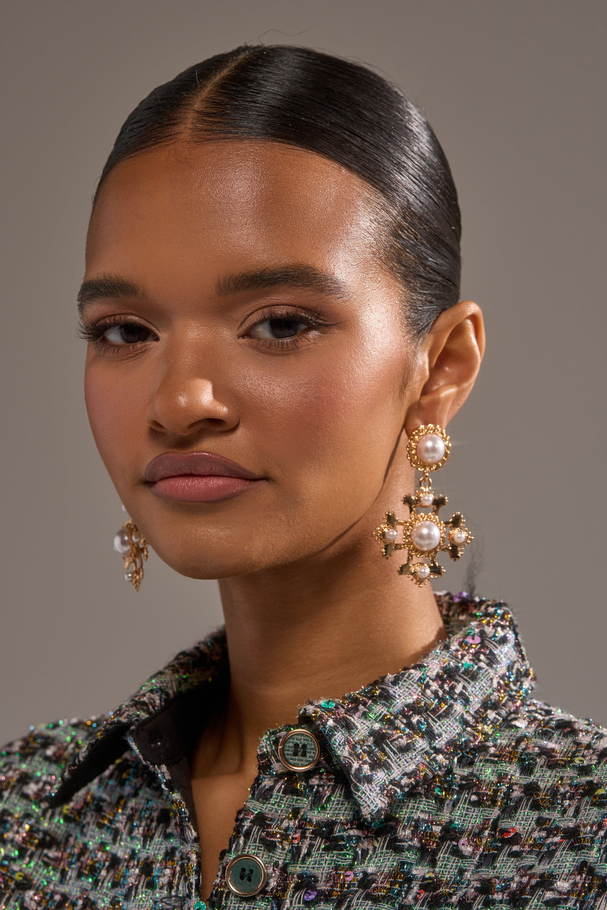 A woman with smooth, dark hair styled back wears the CROSS MY HEART EARRING—large gold and pearl studs—paired with a textured tweed jacket with gold trim, gazing confidently at the camera against a neutral background. 1