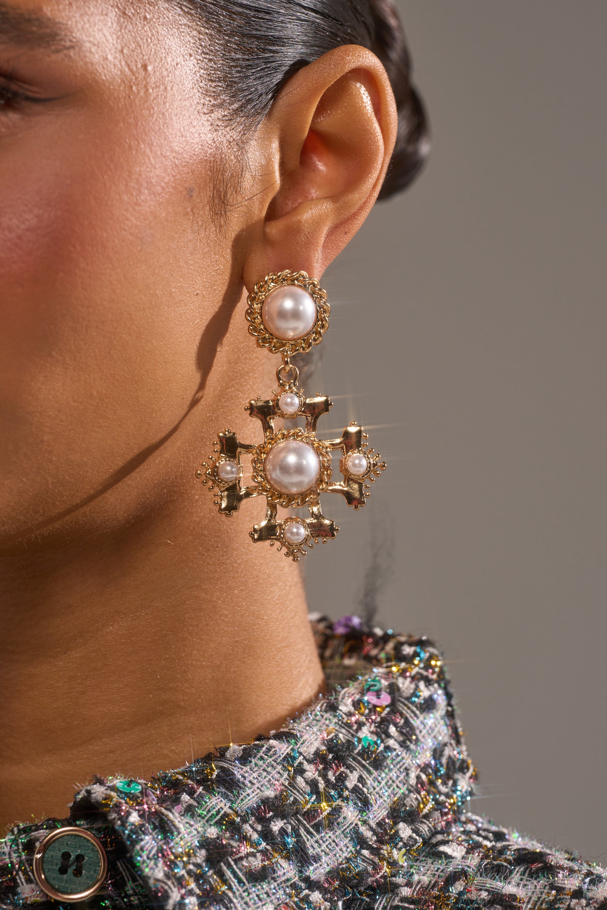 Close-up of a woman's ear wearing the CROSS MY HEART EARRING—an ornate gold piece with large pearls and pearl studs—paired with a textured, multicolored tweed jacket. The background is softly blurred. 2