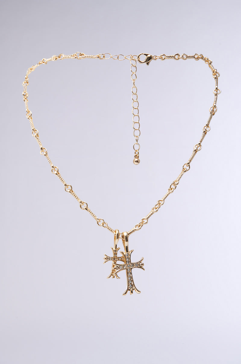 The CROSSES NECKLACE is a faux gold piece with a delicate chain of small links, featuring a rhinestone cross pendant adorned with white stones. It includes an adjustable length, extension chain, and clasp, all on a plain light gray background.