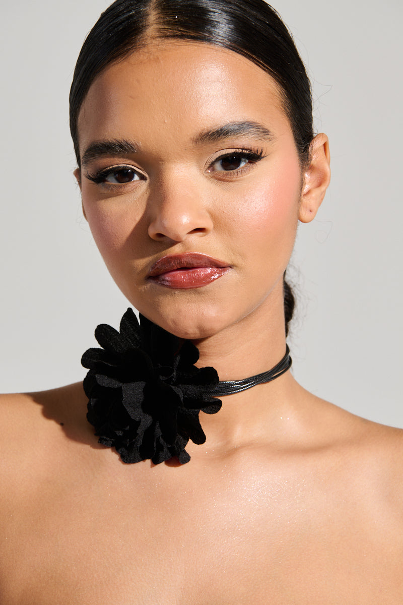 A person with slicked-back hair wears glossy makeup and bold lipstick, showcasing a distinctive accessory: a black choker adorned with the FLOWER DREAMIN CORSAGE NECKLACE, featuring a striking floral centerpiece. The neutral background accentuates their elegant and minimalist appearance. 0