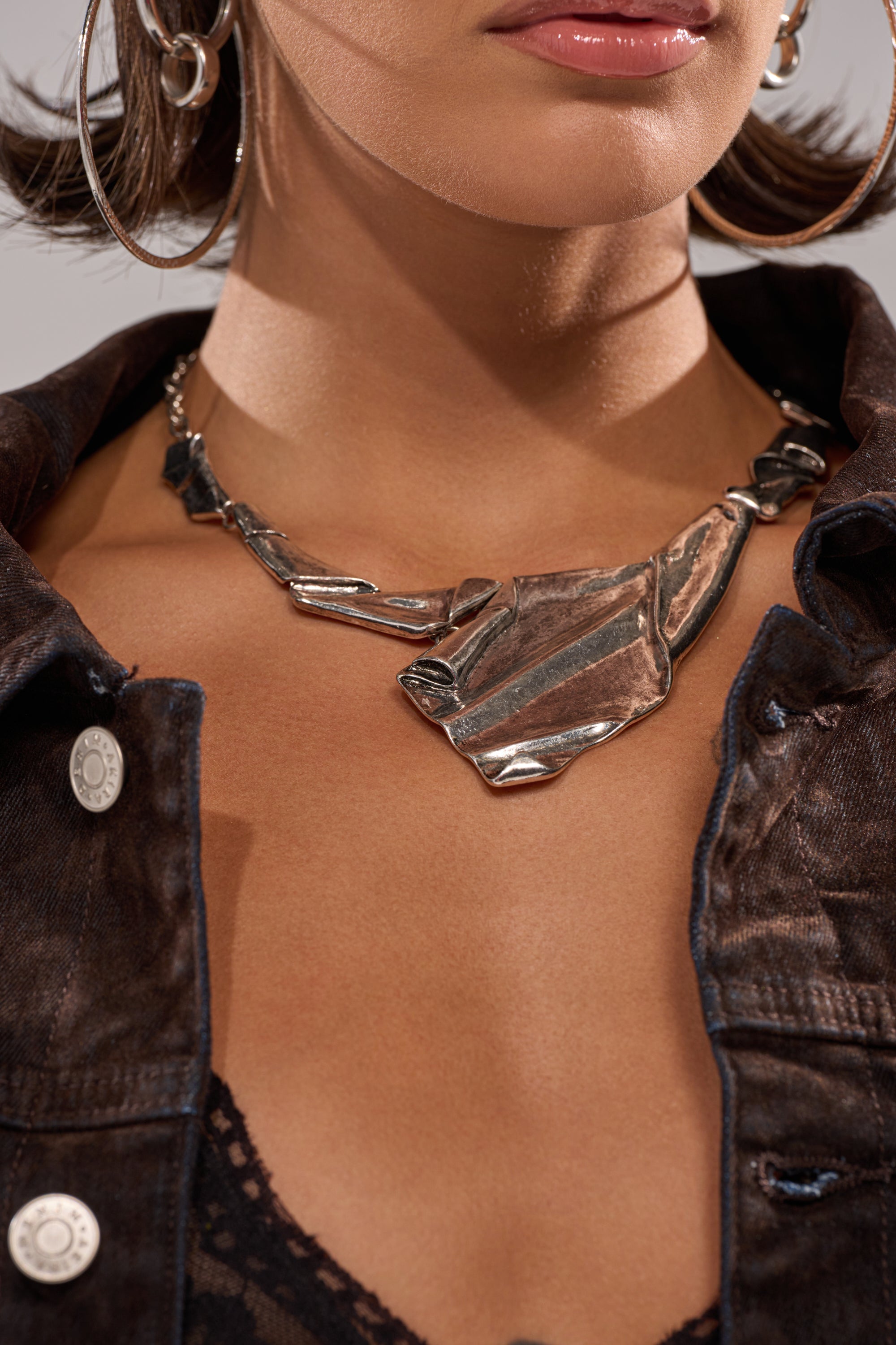 A woman with short hair, glossy lips, and a dark denim jacket over a black lace top wears large silver hoop earrings and the BENT OUTTA SHAPE NECKLACE, its chunky abstract geometric design drawing attention to her jewelry and neckline. 1