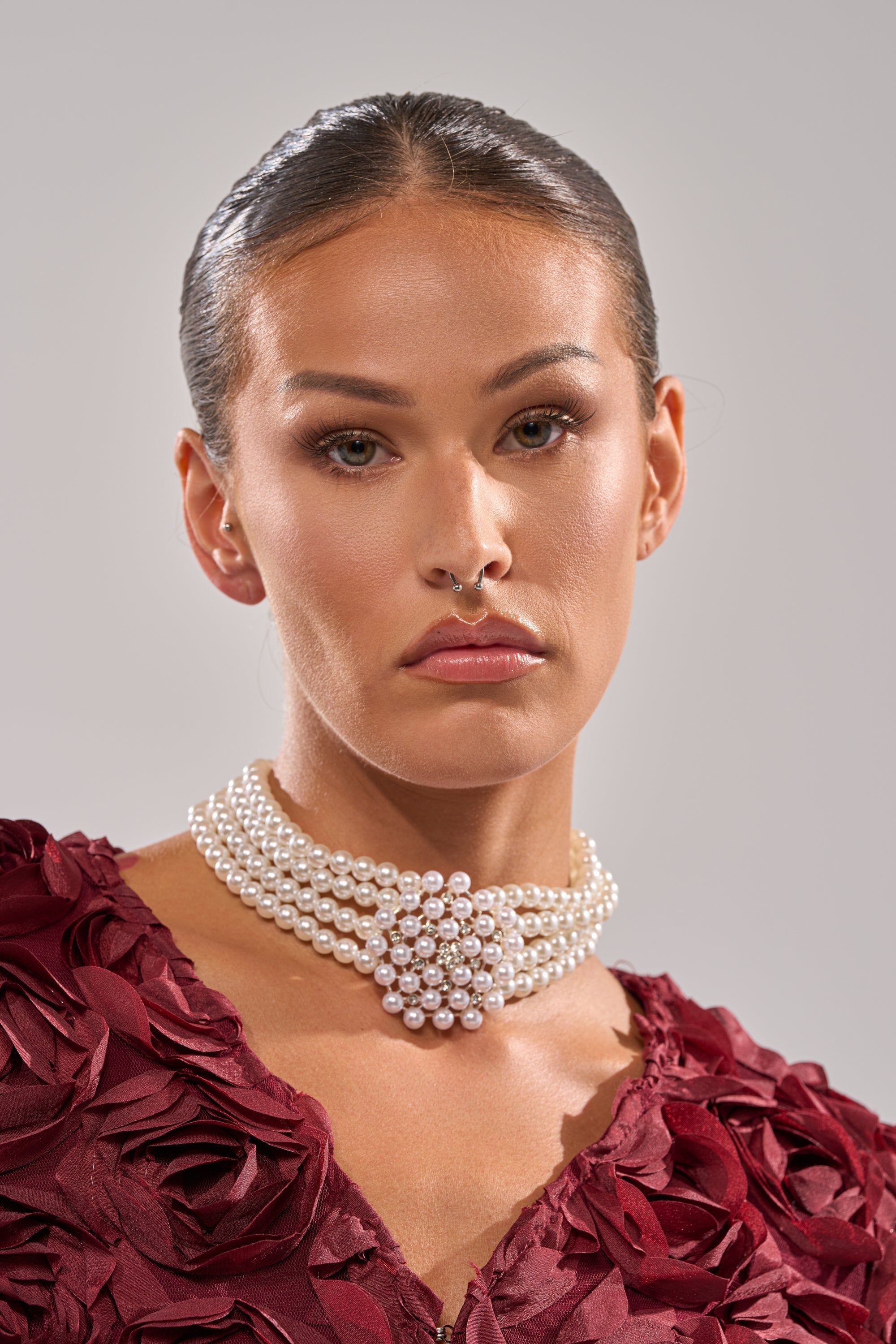 A woman with slicked-back hair wears a deep red textured top and the TRULY PEARL CHOKER—a multi-strand faux pearl necklace with rhinestone details—gazing seriously at the camera against a neutral background. 2