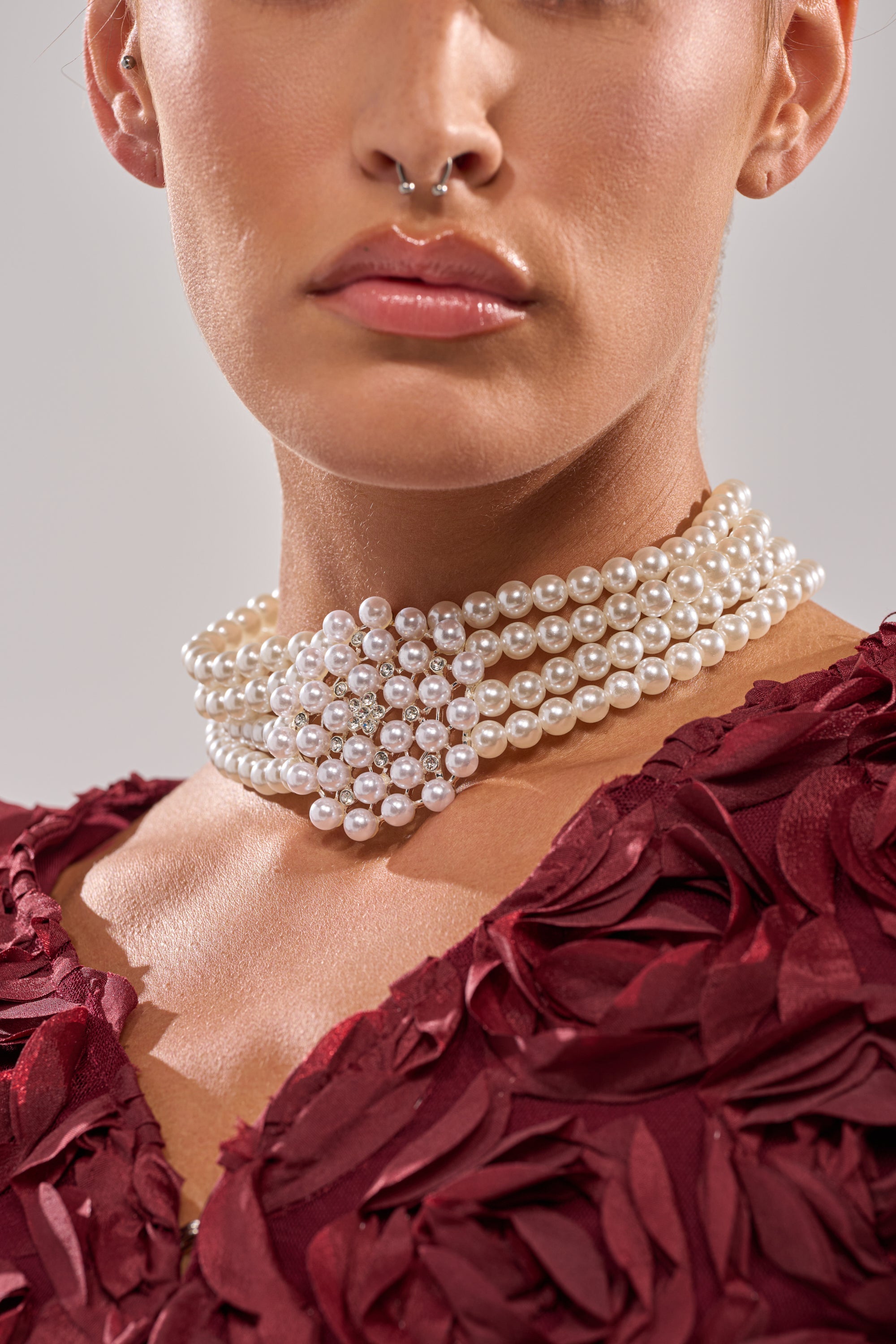 A woman wears the TRULY PEARL CHOKER with rhinestone details, paired with a deep red textured dress and a silver septum ring. The photo highlights her neck and lower face. 3