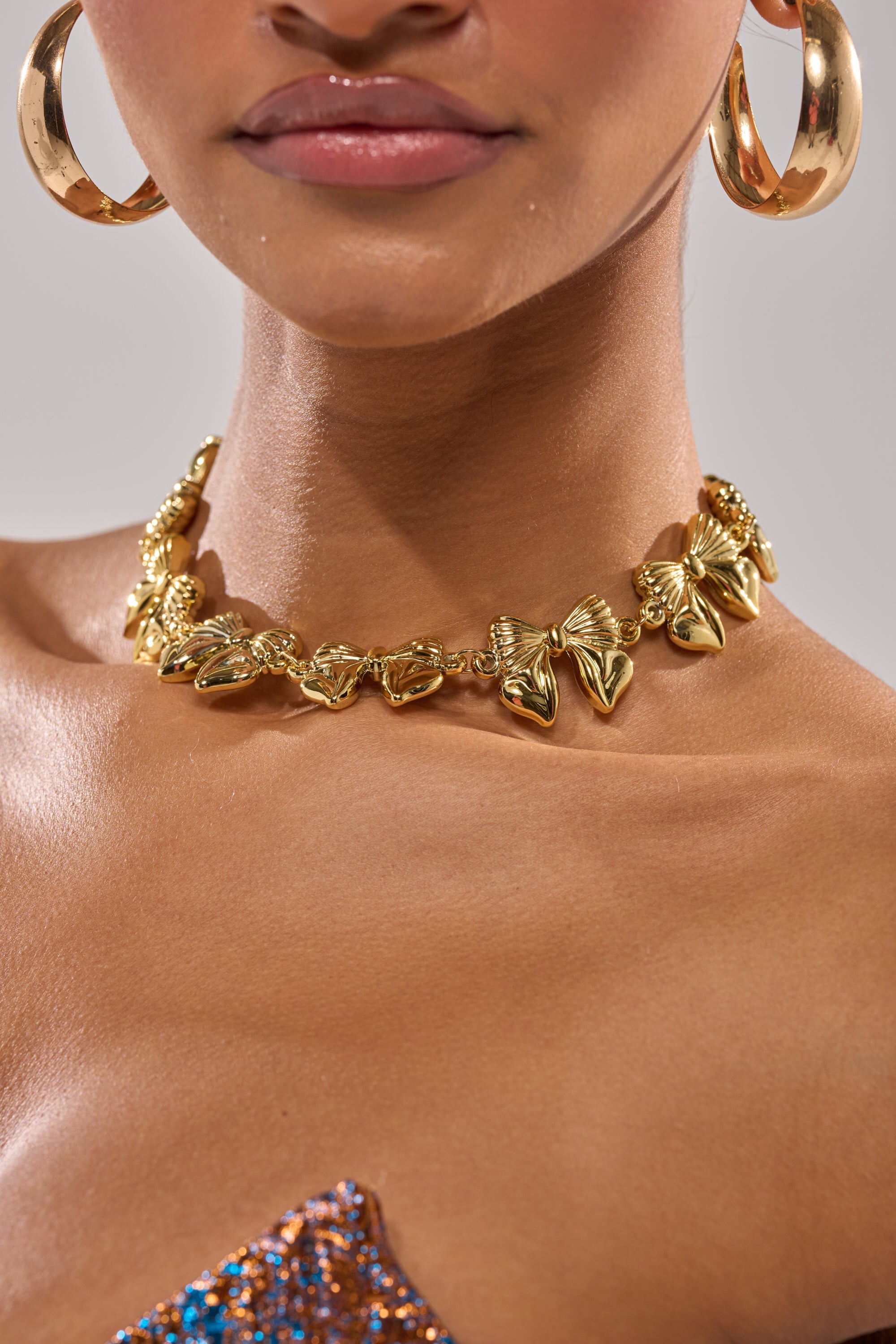 A close-up of a woman wearing large gold hoop earrings and the BOW NECKLACE, a chunky gold-tone piece with butterfly charms and an adjustable clasp, paired with a sparkling strapless top. Her lips are glossy and skin radiant. 1