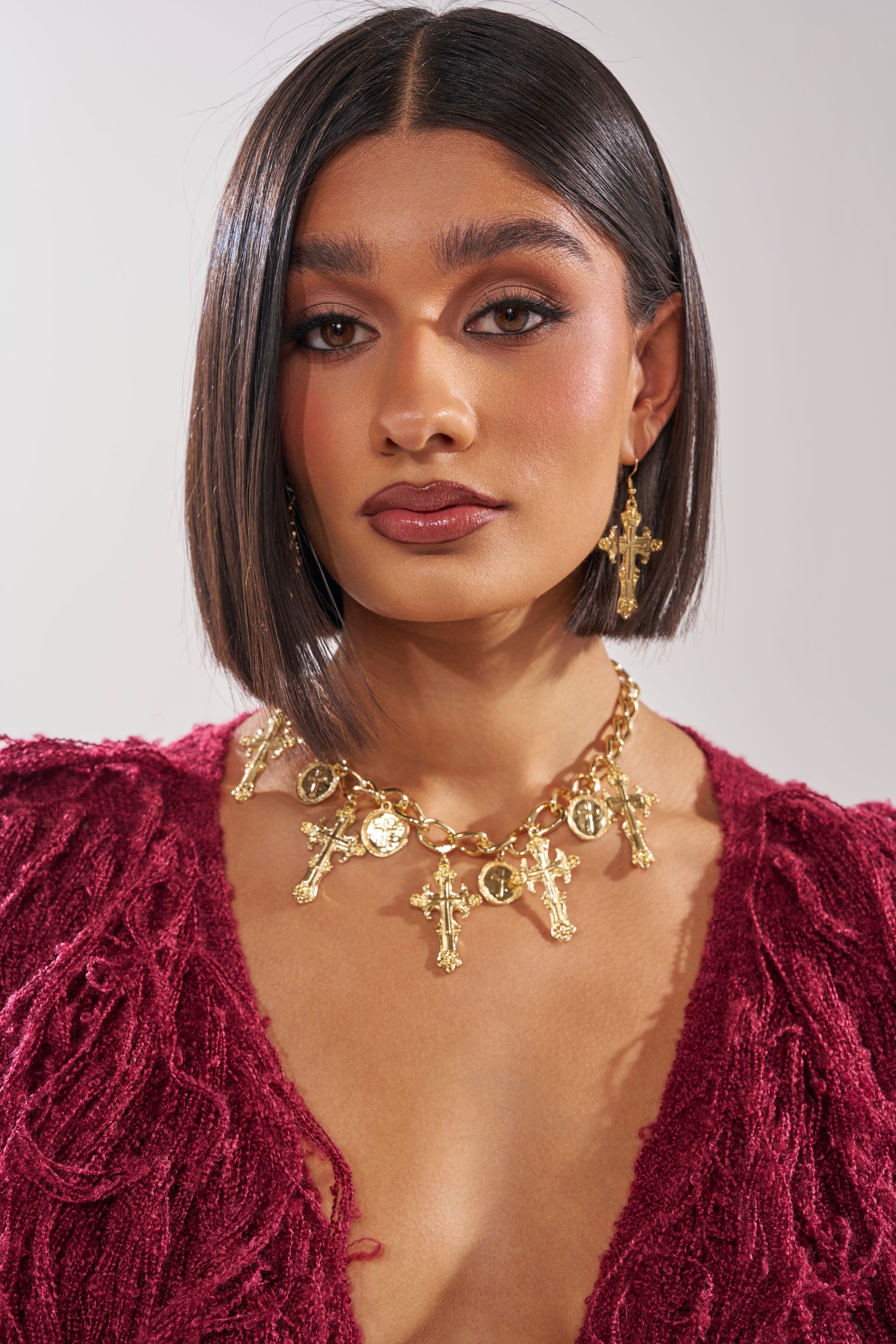 A woman with straight, chin-length dark hair wears a deep red textured top, cross charm earrings, and the CHARMED LIFE NECKLACE—a chunky chain adorned with multiple coin and cross pendants. Her makeup is soft and glowing; her expression, neutral. 9