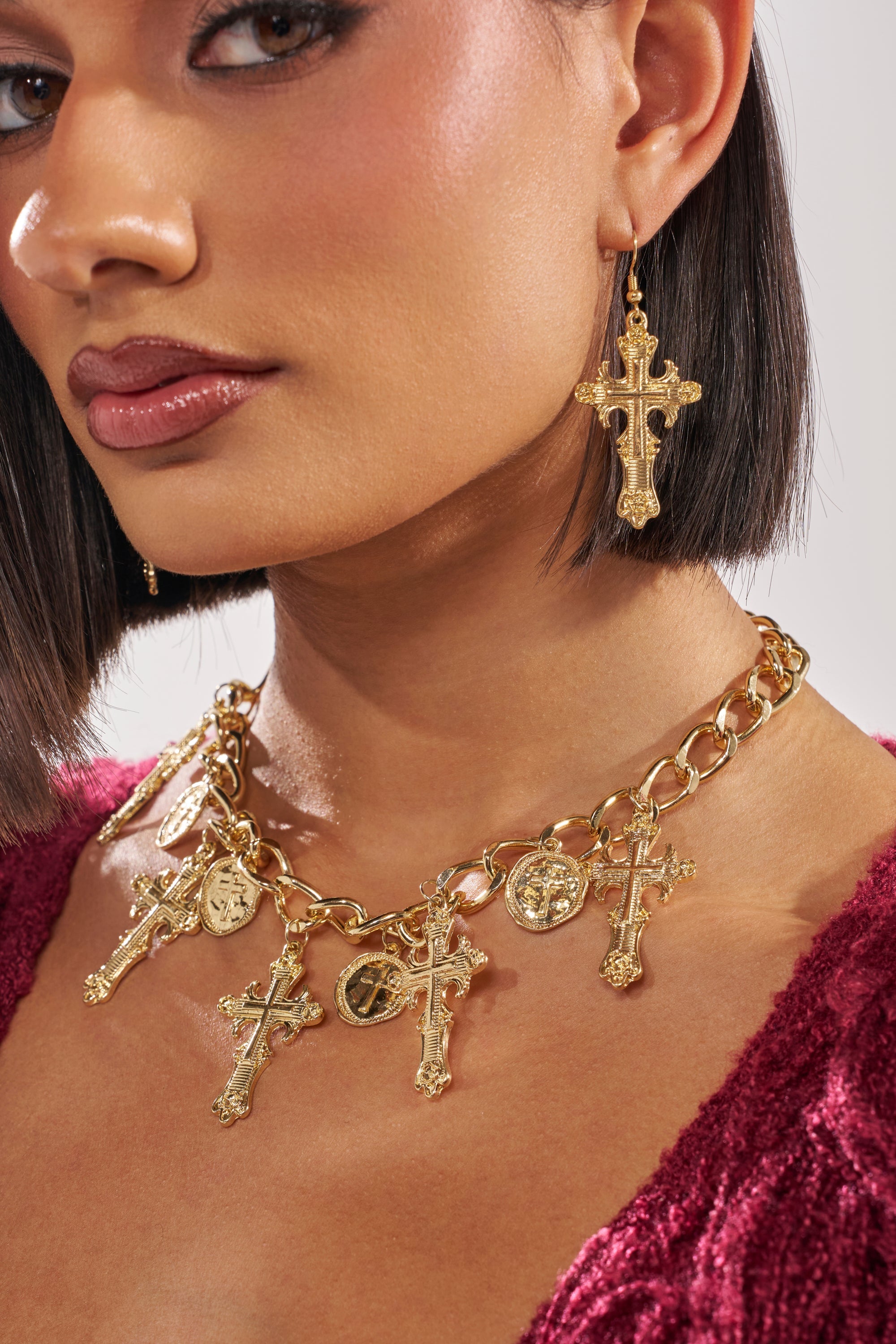 A woman with short dark hair wears a cross earring set and the CHARMED LIFE NECKLACE, a chunky chain jewelry piece adorned with crosses and coins. She pairs it with a deep red textured top. 10