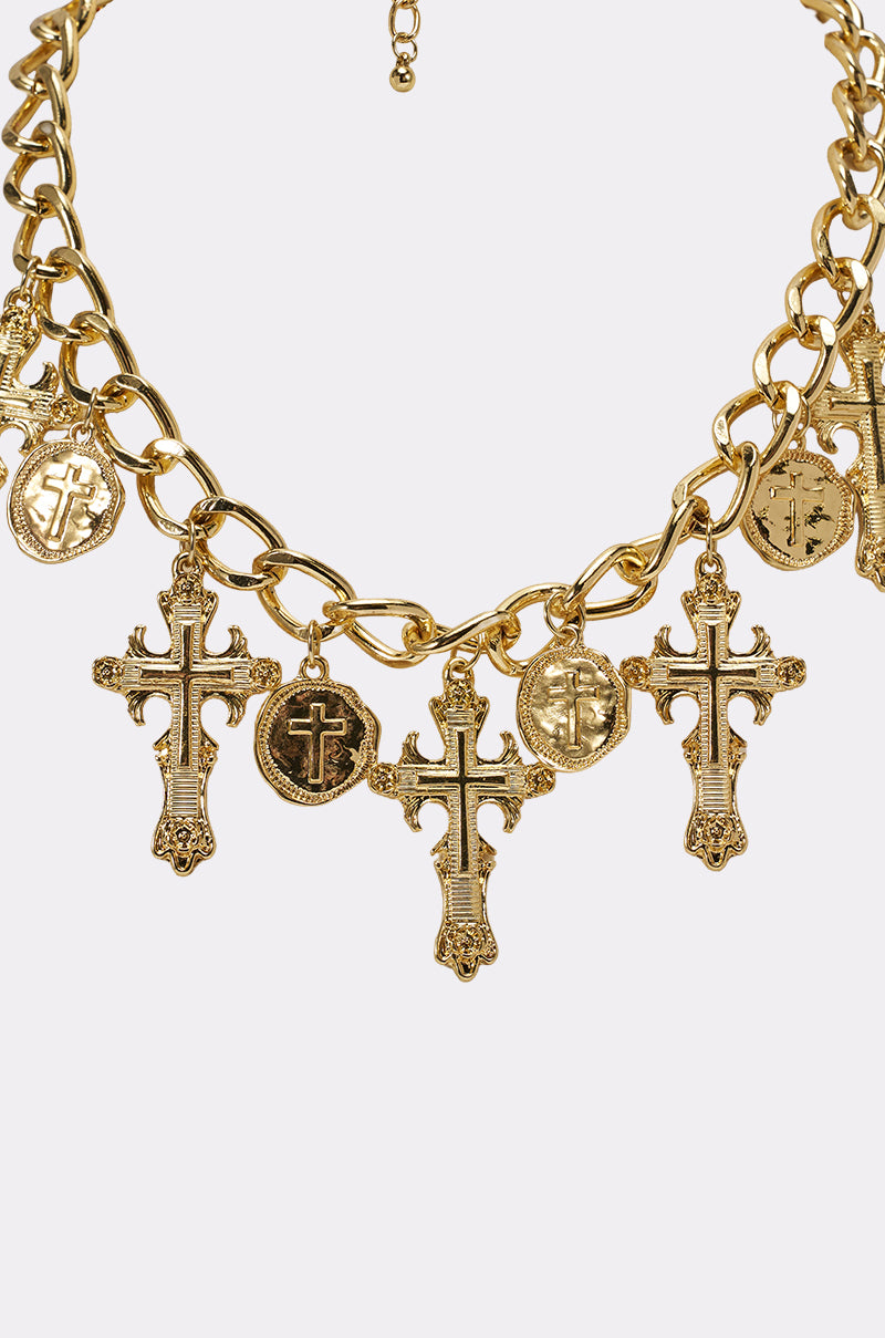 The CHARMED LIFE NECKLACE is a chunky gold chain featuring dangling cross pendants and embossed cross medallion charms, displayed on a plain white background. 7