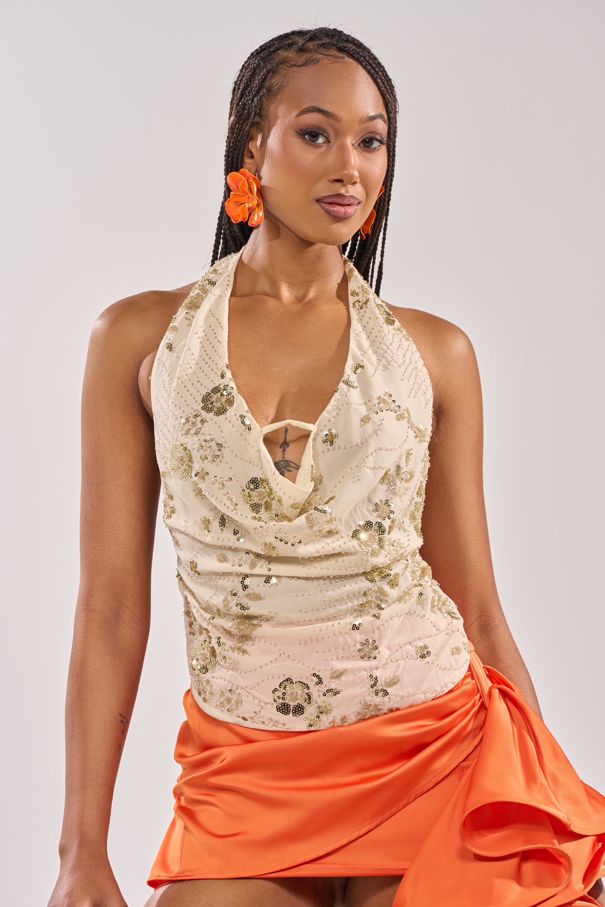 A woman with long braided hair wears orange flower earrings and the JULIE BABY YELLOW HALTER TOP WITH SEQUIN, paired with an orange satin skirt—perfect for festival styling—against a plain light background. 1