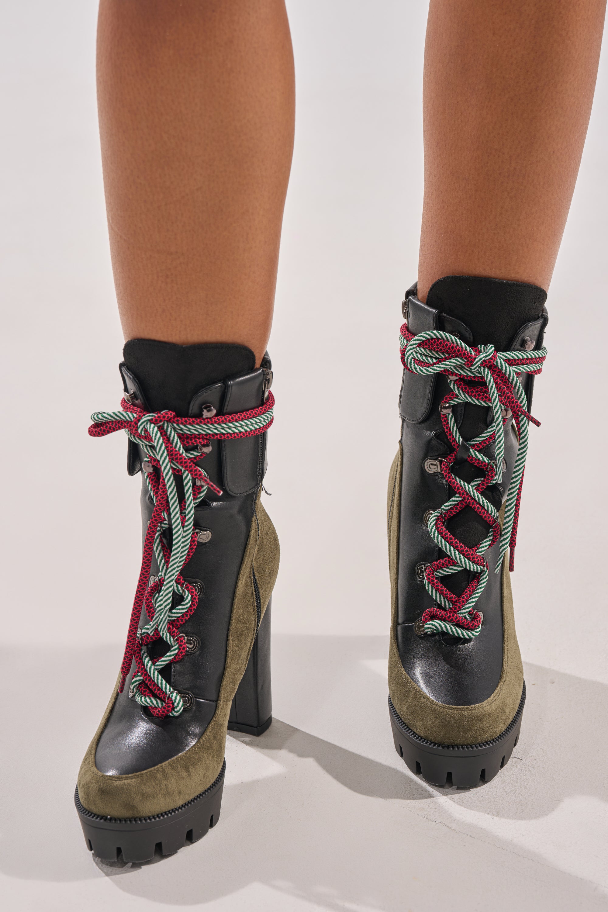 The AKIRA GOLD MUFFLER GREENMULTI CHUNKY HEEL BOOTIE features green faux suede sides, black leather fronts, red-and-green striped laces tied in bows, a platform lug sole, and a chunky heel—shown against a plain light background.