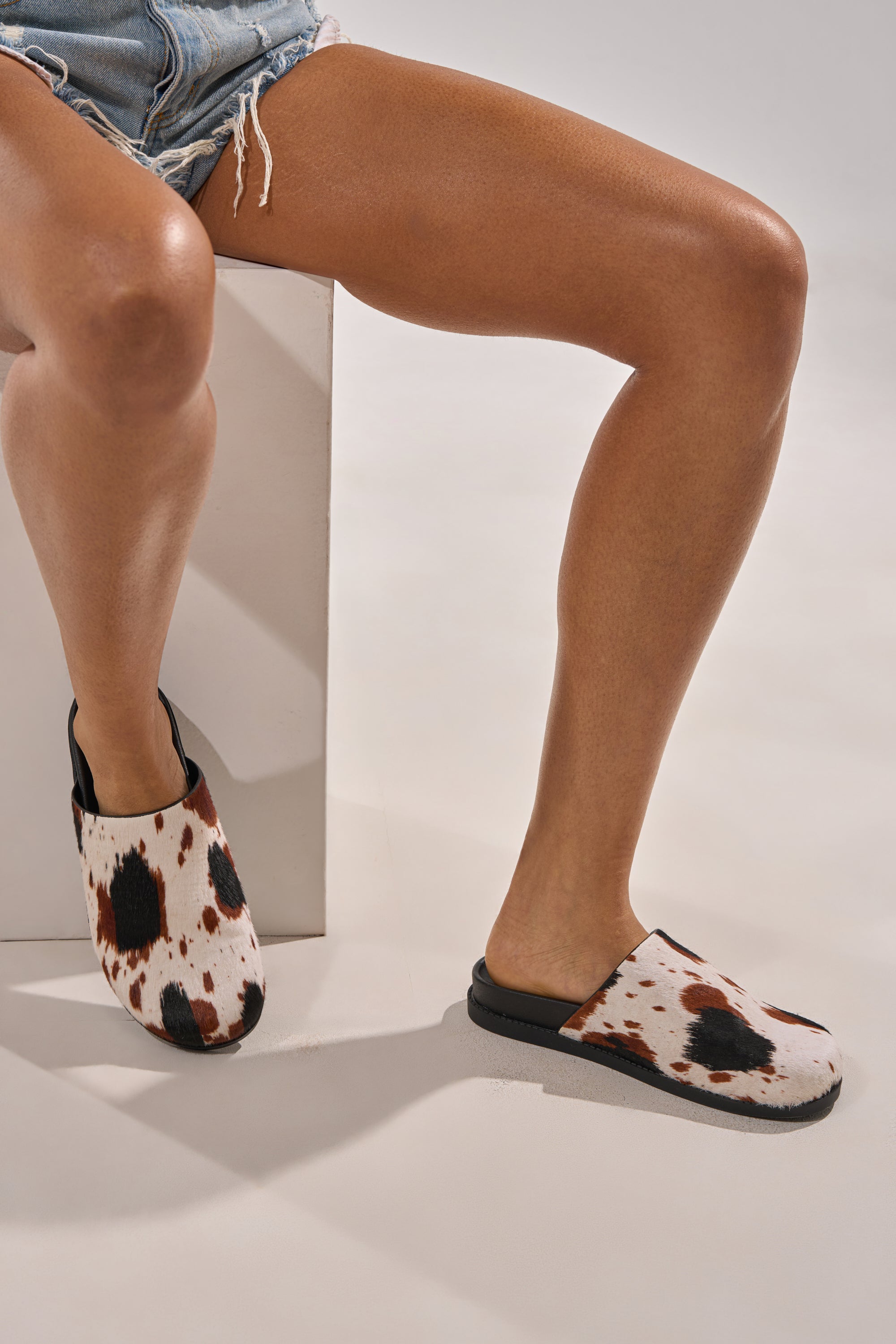 Modeling the AZALEA WANG MUKEY COW PRINT CALF HAIR FLAT, a person in distressed denim shorts sits on a white block wearing genuine calf hair cow print flats with black soles, set against a neutral background. 1