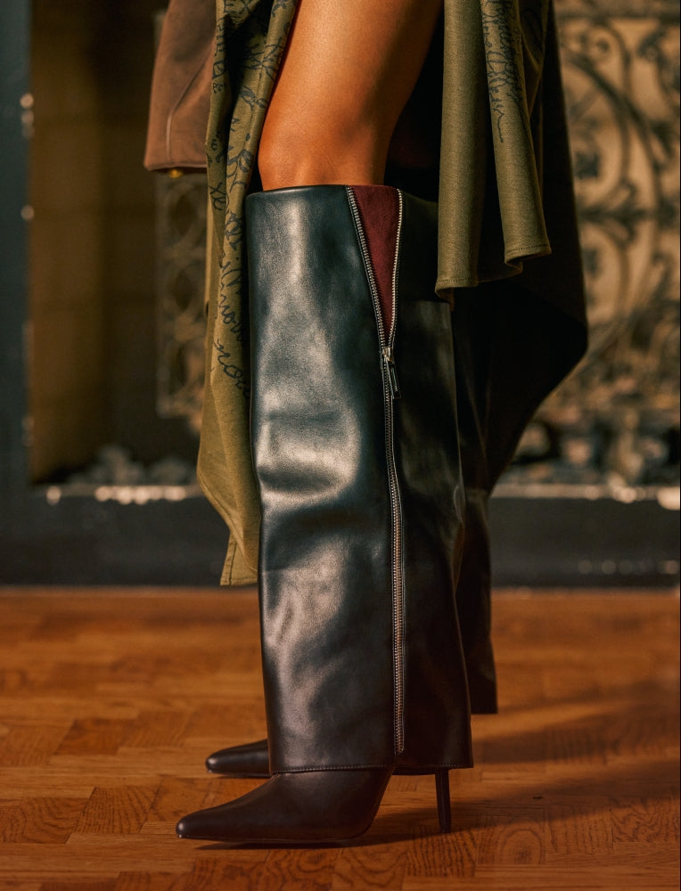 A close-up of a person wearing a tall black leather boot with a side zipper, shown from the lower torso down on a wooden floor.