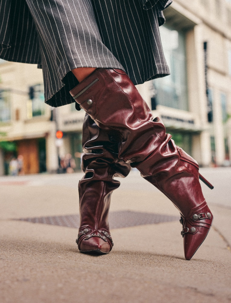 A close-up of a person wearing knee-high burgundy leather slouchy boots with pointed toes and silver buckle details, walking on a city street.