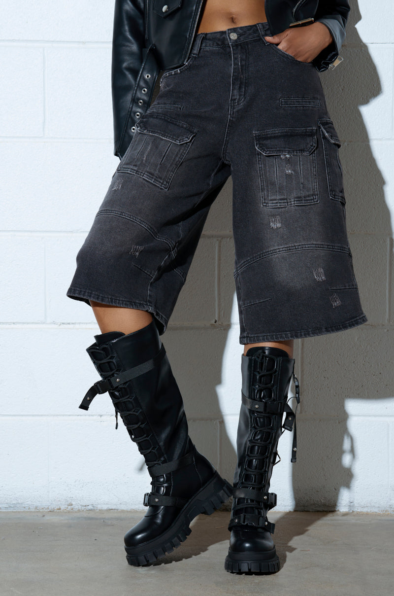 A person wearing AZALEA WANG MYRON BLACK BOOT with thick soles and laces, paired with loose-fitting dark denim cargo shorts and a black leather jacket, stands against a plain white wall.
