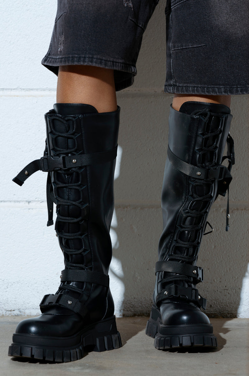 Person standing against a light-colored wall, wearing the AZALEA WANG MYRON BLACK BOOT, knee-high with thick soles and paired with black shorts.